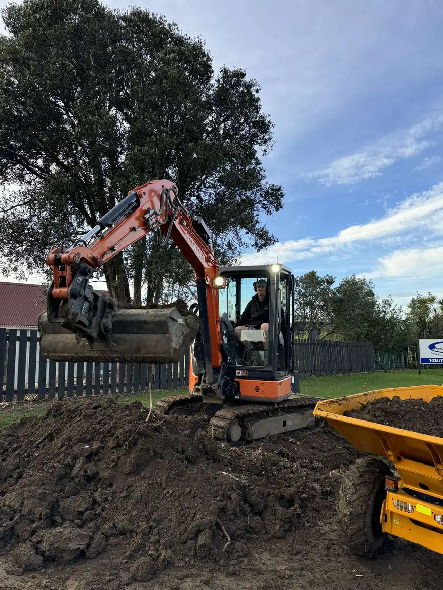 Excavator lifting a bucket of soil beside a yellow tipper truck on a prepared site
