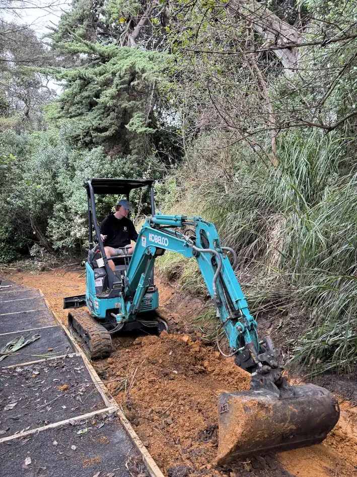 Small blue mini excavator digging orange clay on a sloping bush site beside a path